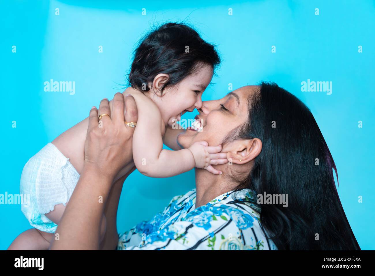 Smiling indian mother playing with her six months cute little baby in diaper isolated over blue background. Happy family. Asian mom with infant child Stock Photo