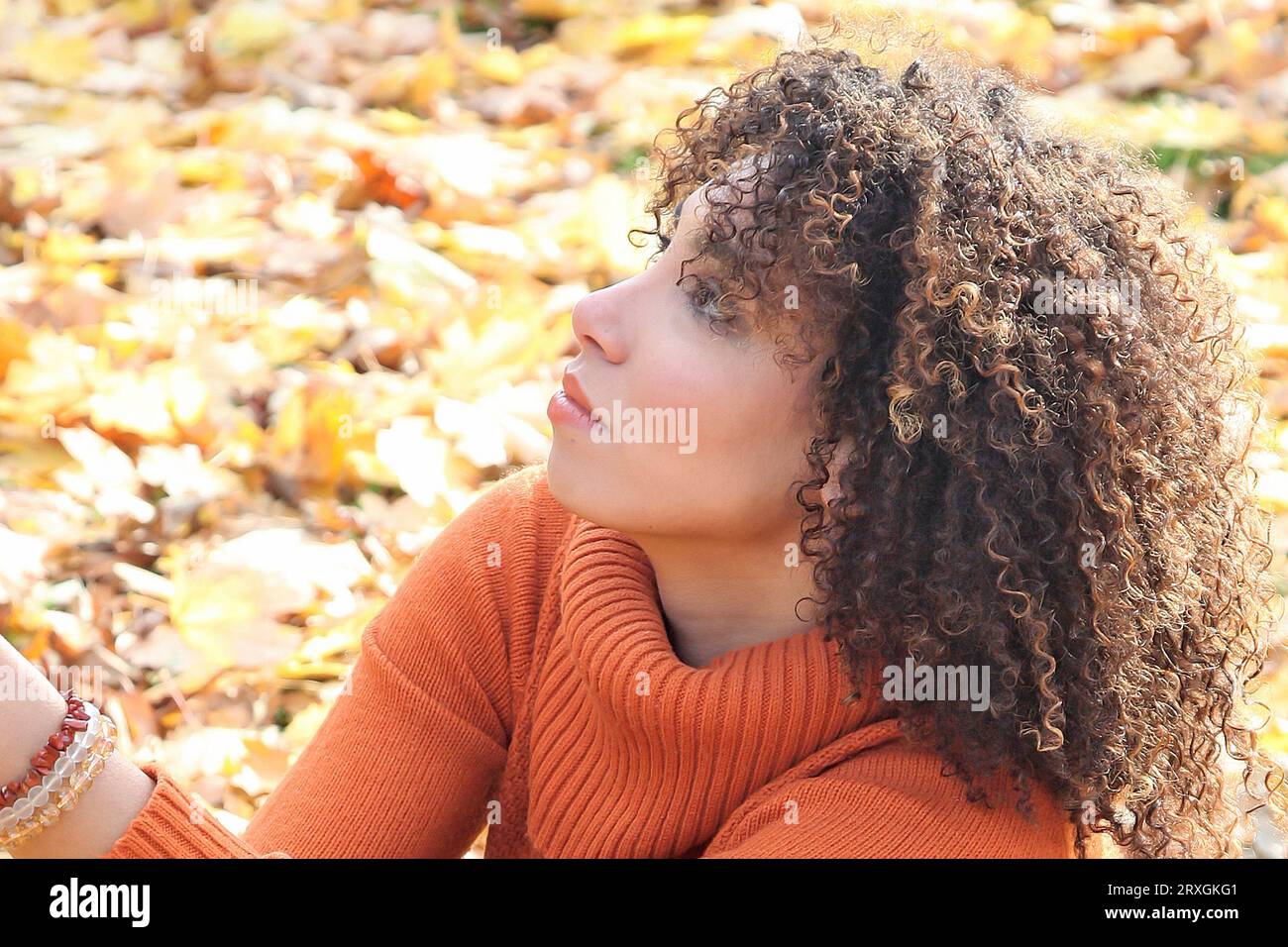 Curly haired woman with orange sweater posing with the autumn leaves in background Stock Photo