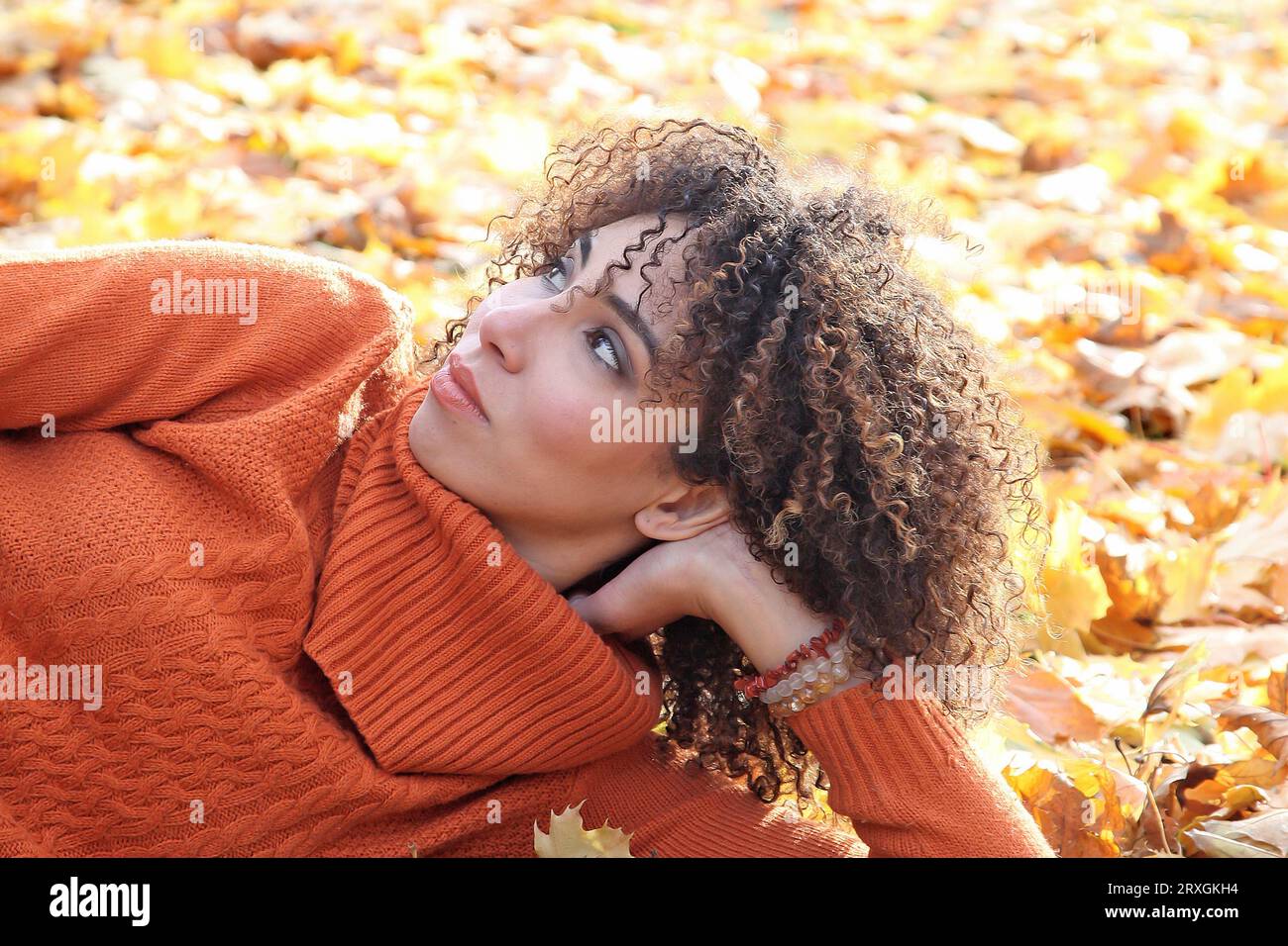 Curly haired woman with orange sweater posing with the autumn leaves in background Stock Photo