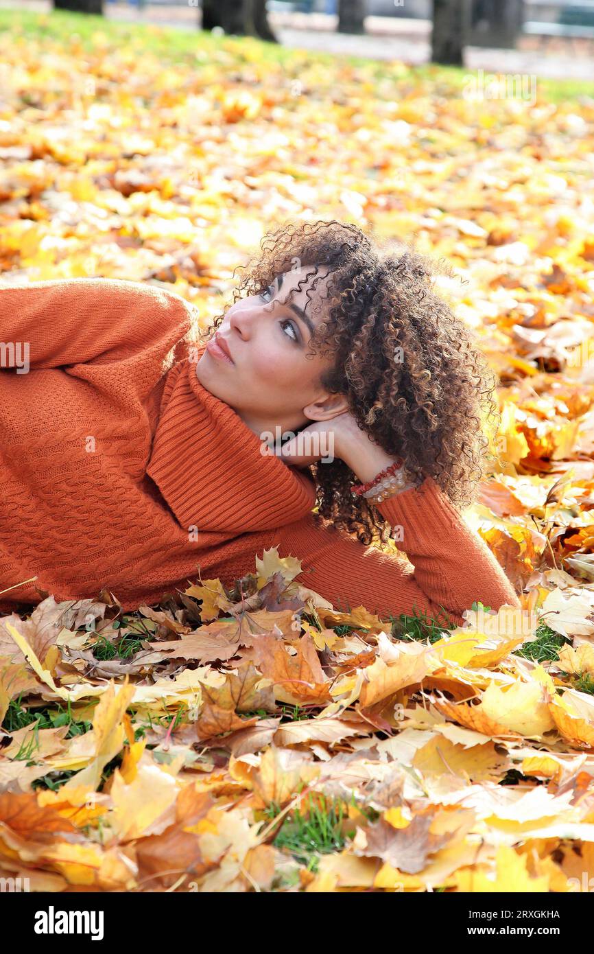 Curly haired woman with orange sweater posing with the autumn leaves in background Stock Photo