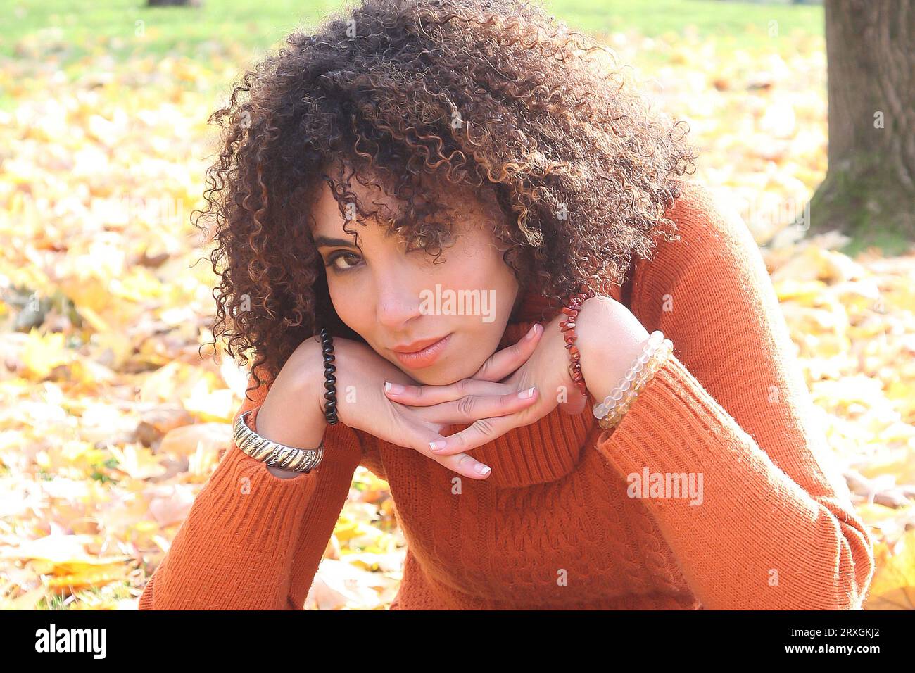 Curly haired woman with orange sweater posing with the autumn leaves in background Stock Photo