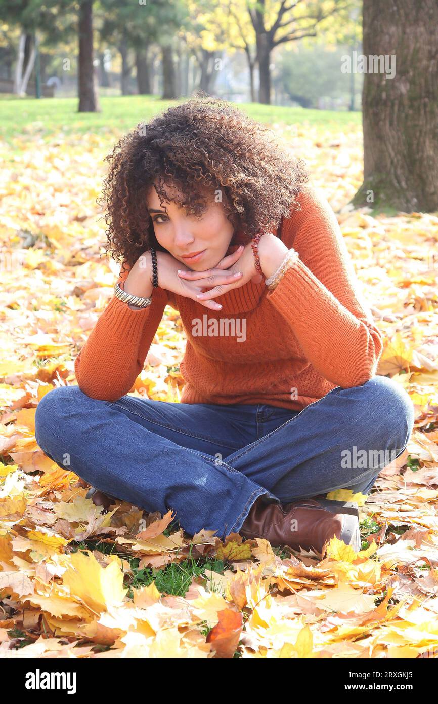 Curly haired woman with orange sweater posing with the autumn leaves in background Stock Photo