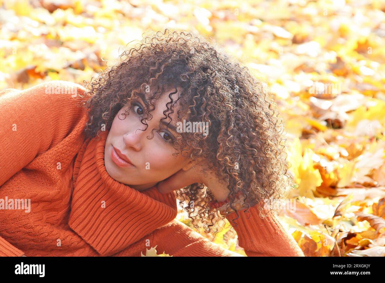 Curly haired woman with orange sweater posing with the autumn leaves in background Stock Photo