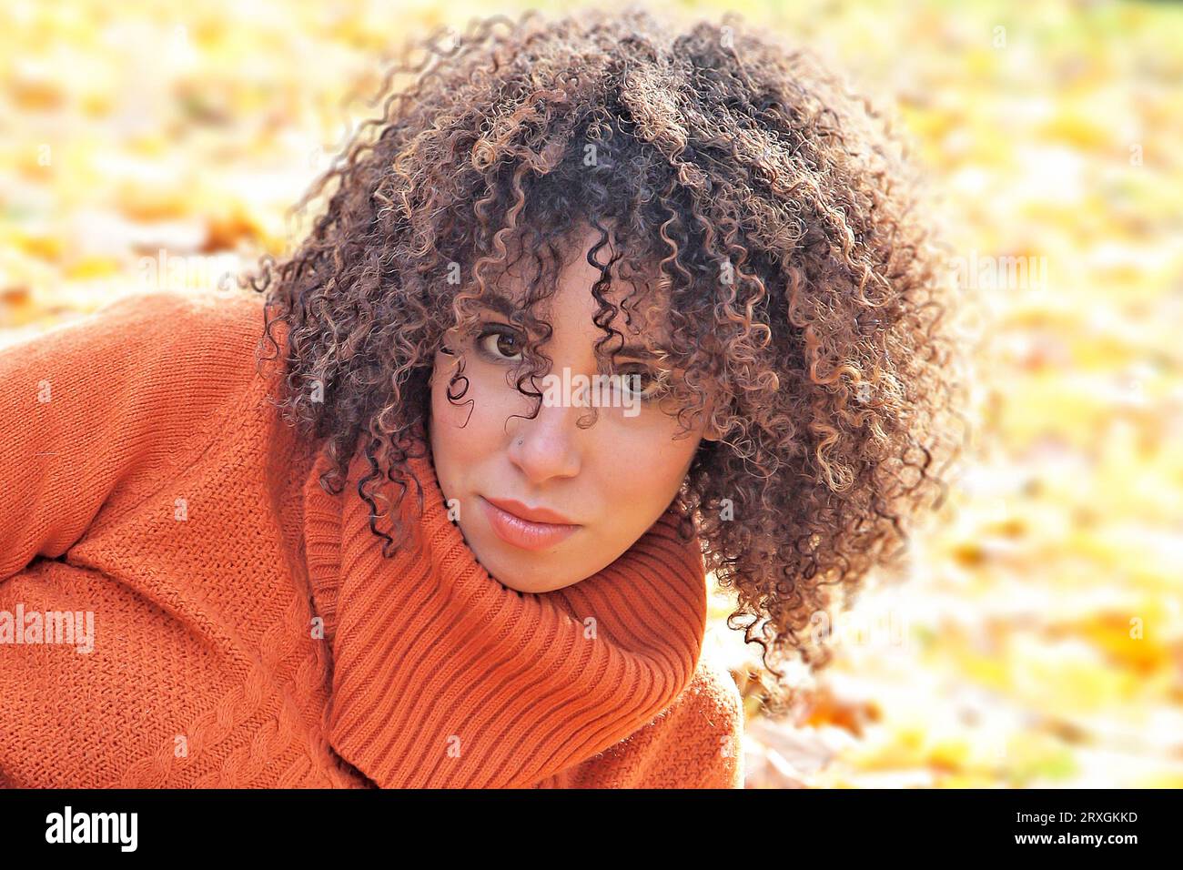 Curly haired woman with orange sweater posing with the autumn leaves in background Stock Photo