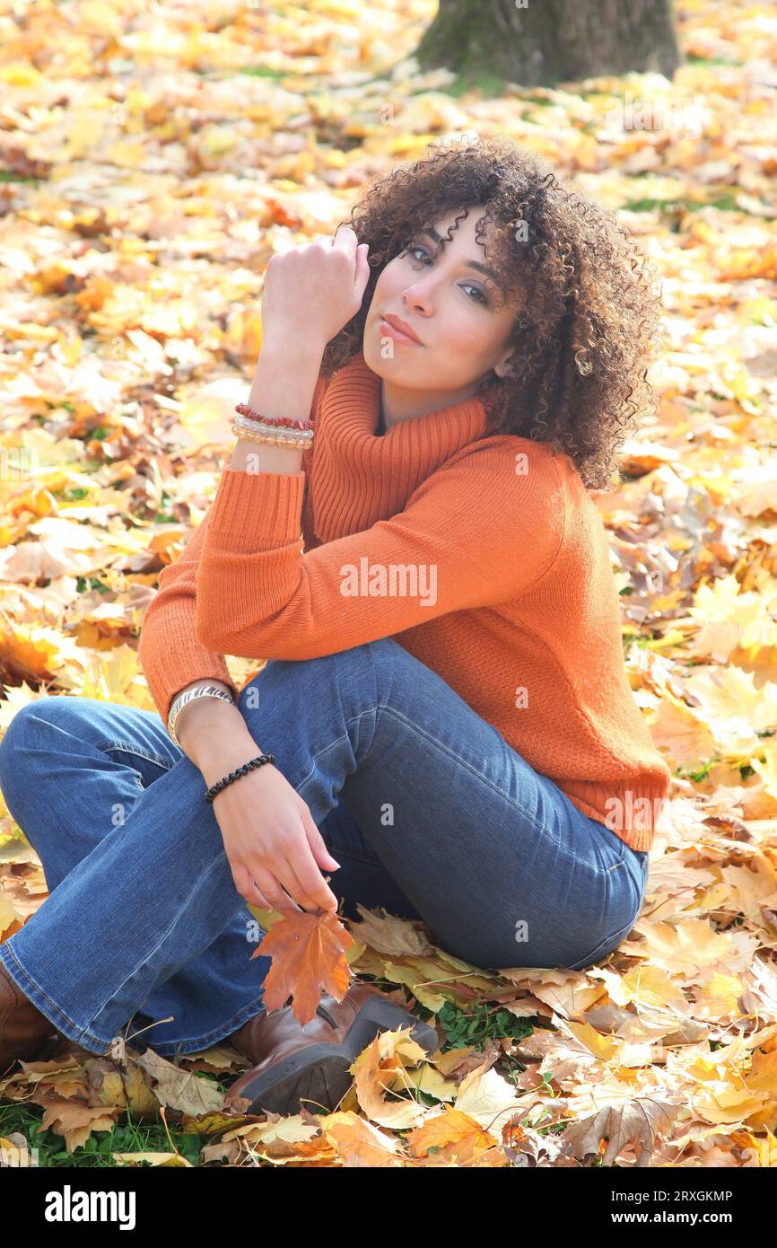 Curly haired woman with orange sweater posing with the autumn leaves in background Stock Photo