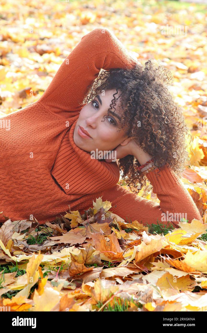 Curly haired woman with orange sweater posing with the autumn leaves in background Stock Photo