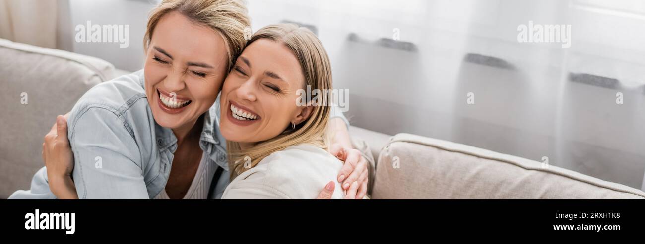 contented lovely sisters in casual outfits sitting on sofa hugging and smiling, bonding, banner Stock Photo
