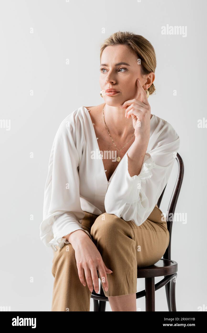 beautiful blonde woman in formal wear sitting on chair and touching her face, style and fashion Stock Photo