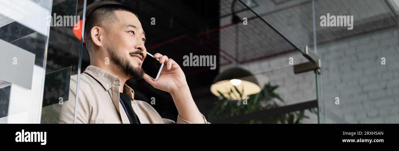 happy asian man having business phone call on smartphone near glass door in office, banner Stock Photo