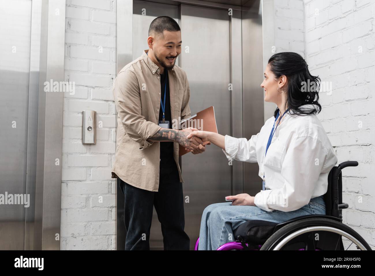 inclusion and diversity, cheerful asian man shaking hands with disabled woman near office elevators Stock Photo