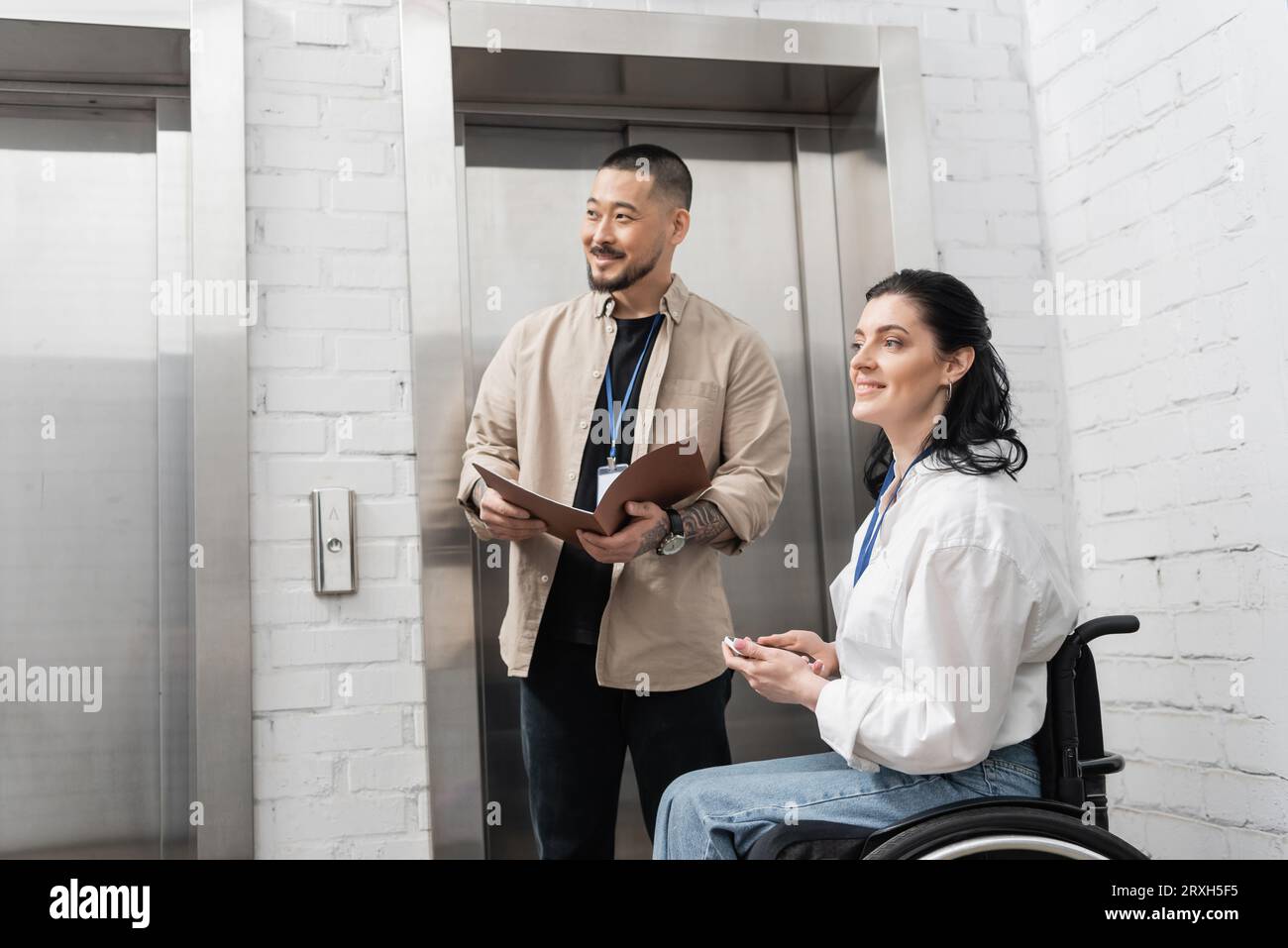 inclusion and diversity, happy asian man folder near disabled woman and office elevators, look away Stock Photo