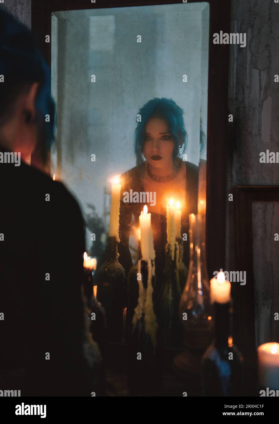 Indoors portrait of lovely goth girl among candles. Blue-haired gothic lady looking into the old dirty mirror. Young witch. Vintage style Stock Photo