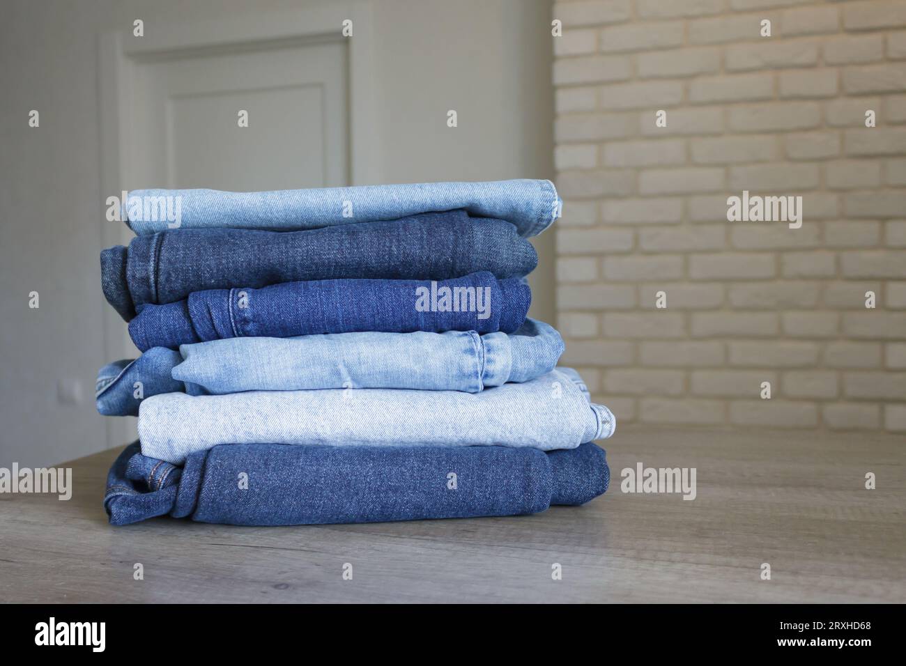 A neatly folded stack of clothes - jeans in different shades of blue on a wooden table against the backdrop of a bright room. Stock Photo