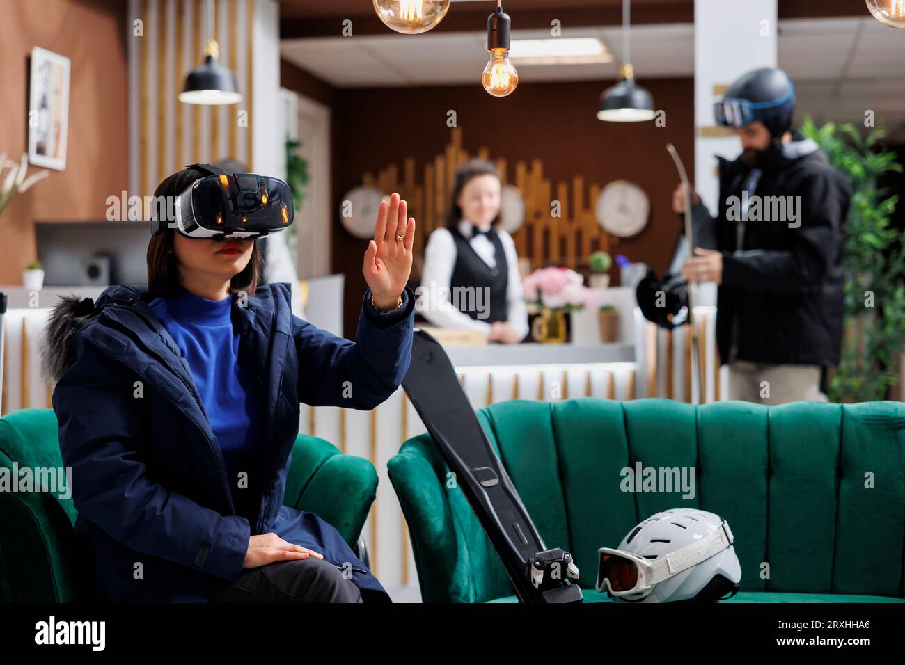 Asian lady wearing digital VR headset having skiing equipment and winter jacket in hotel lobby. Female tourist searching with vr glasses for best slopes at ski mountain resort snow adventure. Stock Photo