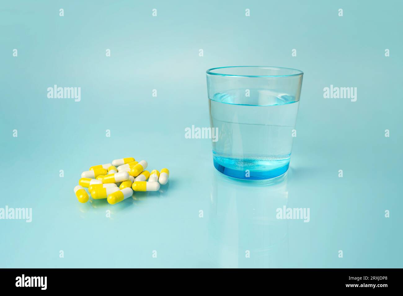 white and yellow medical pills, vitamins, in the distance there is a beautiful blue glass with clean water, on a harmonious bright blue background. Ta Stock Photo