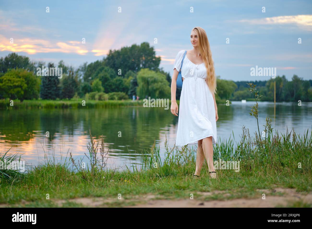Young woman in long white sundress standing on the river bank during sunset Stock Photo