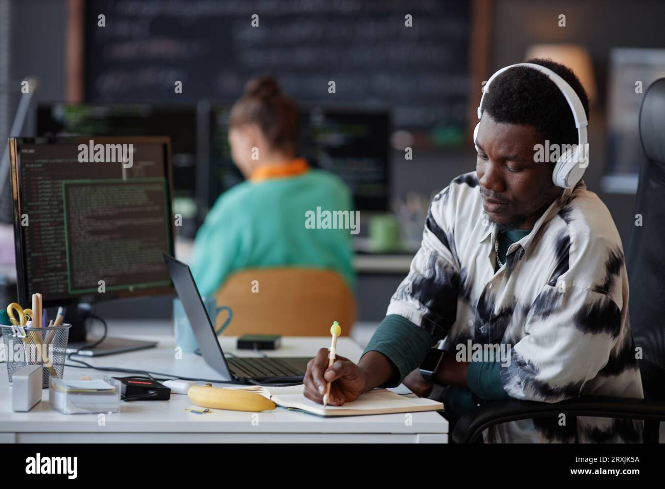 Serious programmer in headphones making notes in notebook while working with security codes on computer at his workplace Stock Photo