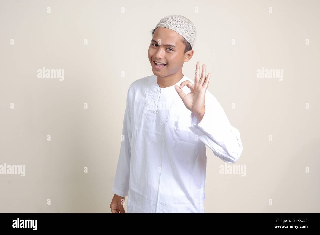Portrait of attractive Asian muslim man in white shirt with skullcap showing ok hand gesture and smiling looking at camera. Advertising concept. Isola Stock Photo