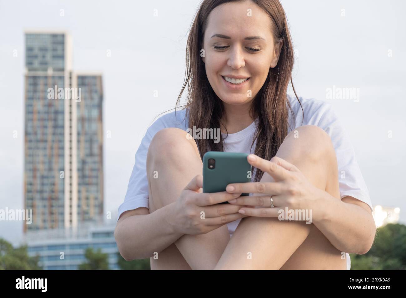Smiling young Caucasian woman using applications on mobile phone, texting message on smartphone. Happy girl checking mobile apps outdoors using Stock Photo