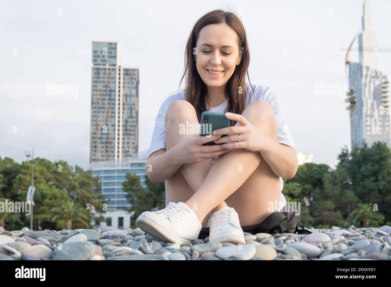 Young happy smiling pretty woman using applications on mobile phone, texting message on smartphone. Cheerful girl checking mobile apps outdoors using Stock Photo