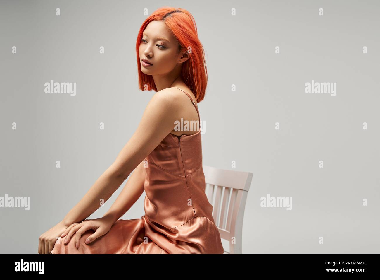elegant asian woman sitting on chair in pastel pink silk slip dress on grey background, serenity Stock Photo