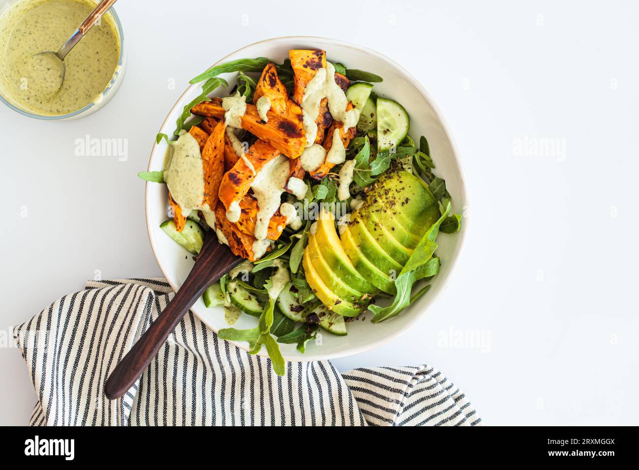 Baked sweet potato wedges salad with cucumber, avocado and creamy tahini dressing, white background, top view. Vegan recipe concept. Stock Photo