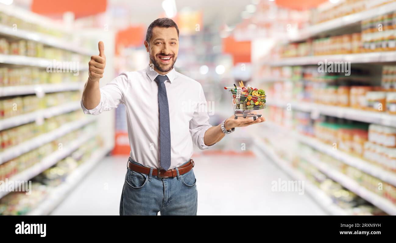Young smiling man with a small shopping cart on his palm gesturing thumbs up in a supermarket Stock Photo