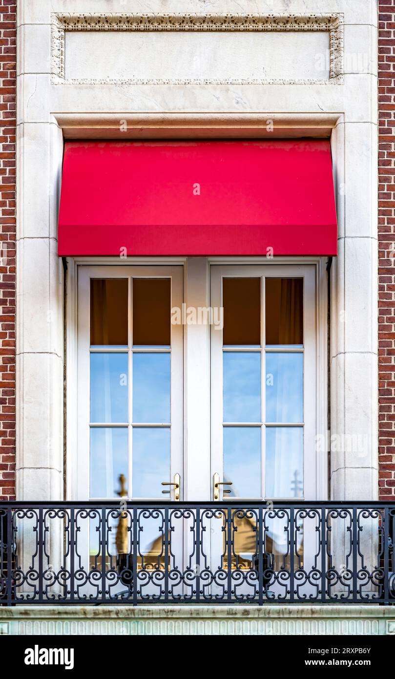 Tall double doors with glass and a bright red textile canopy open onto a small balcony with wrought iron tracery railings in a classic brick building Stock Photo
