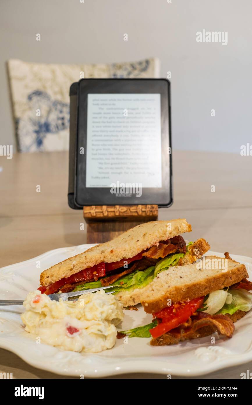 A meal while reading a book on a Kindle, eating a BLT, a bacon, tomato & lettuce sandwich made with grained bread & served on a white plate. USA. Stock Photo