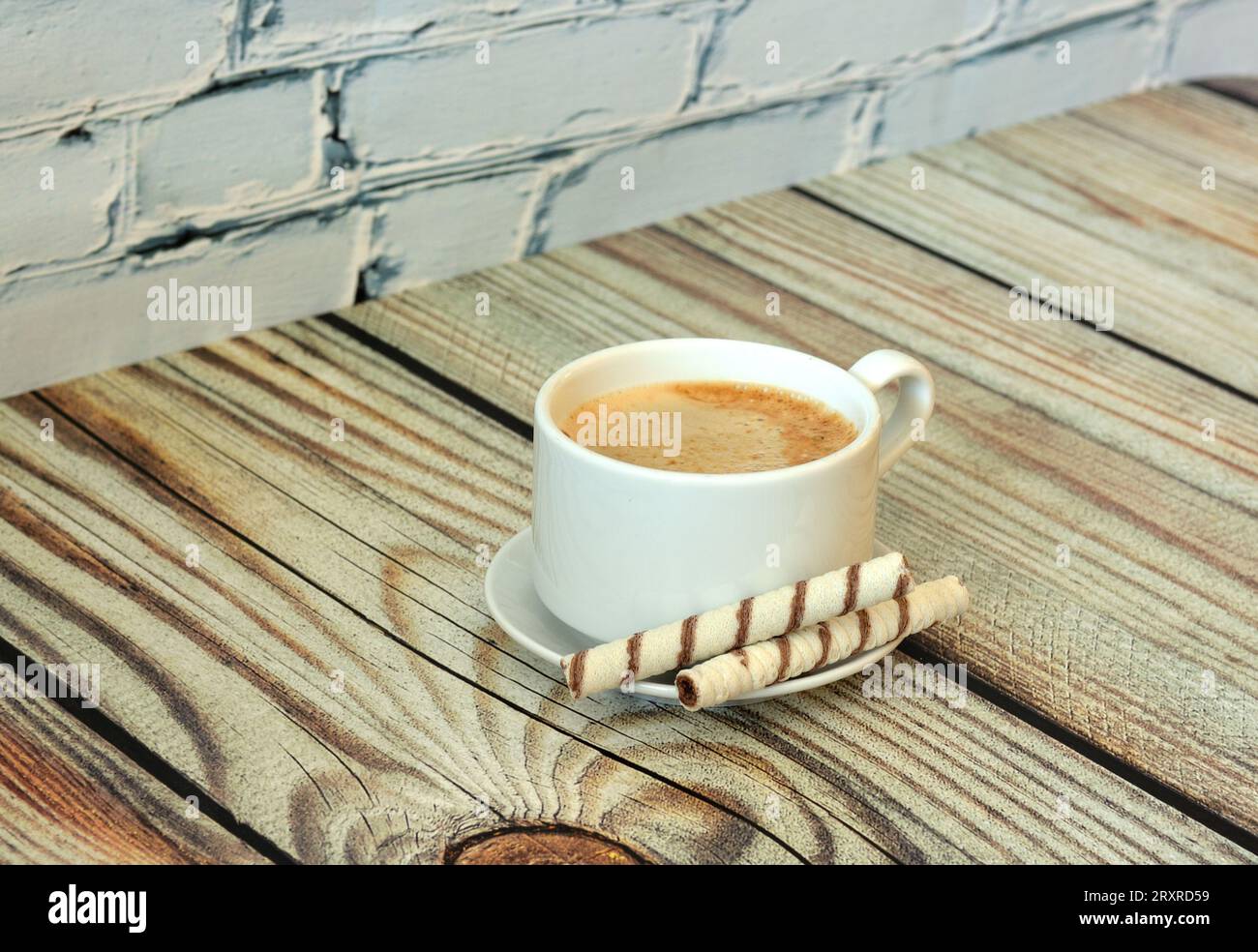 A cup of fresh cappuccino on a saucer with two wafer rolls on a light wooden table. Close-up. Stock Photo