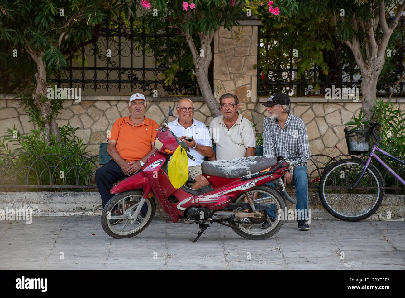 a group of elderly retired greek gentlemen sitting on a bench in zante town watching the world go by with a moped and a bicycle parked next to them. Stock Photo