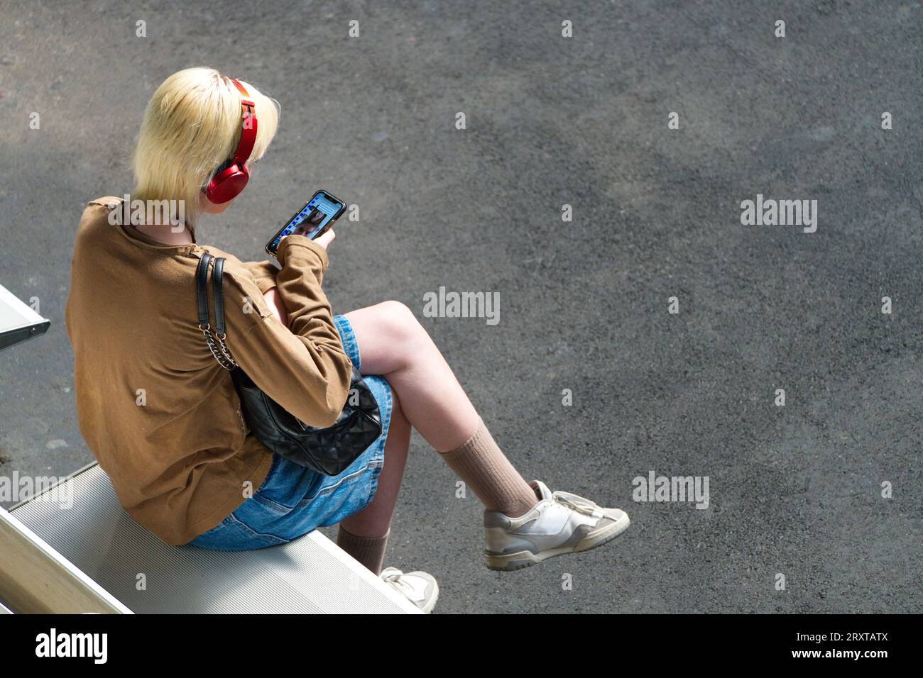 Young person looking at phone wearing headphones and sneakers sitting on seat using social media, in Melbourne city, Australia. Stock Photo