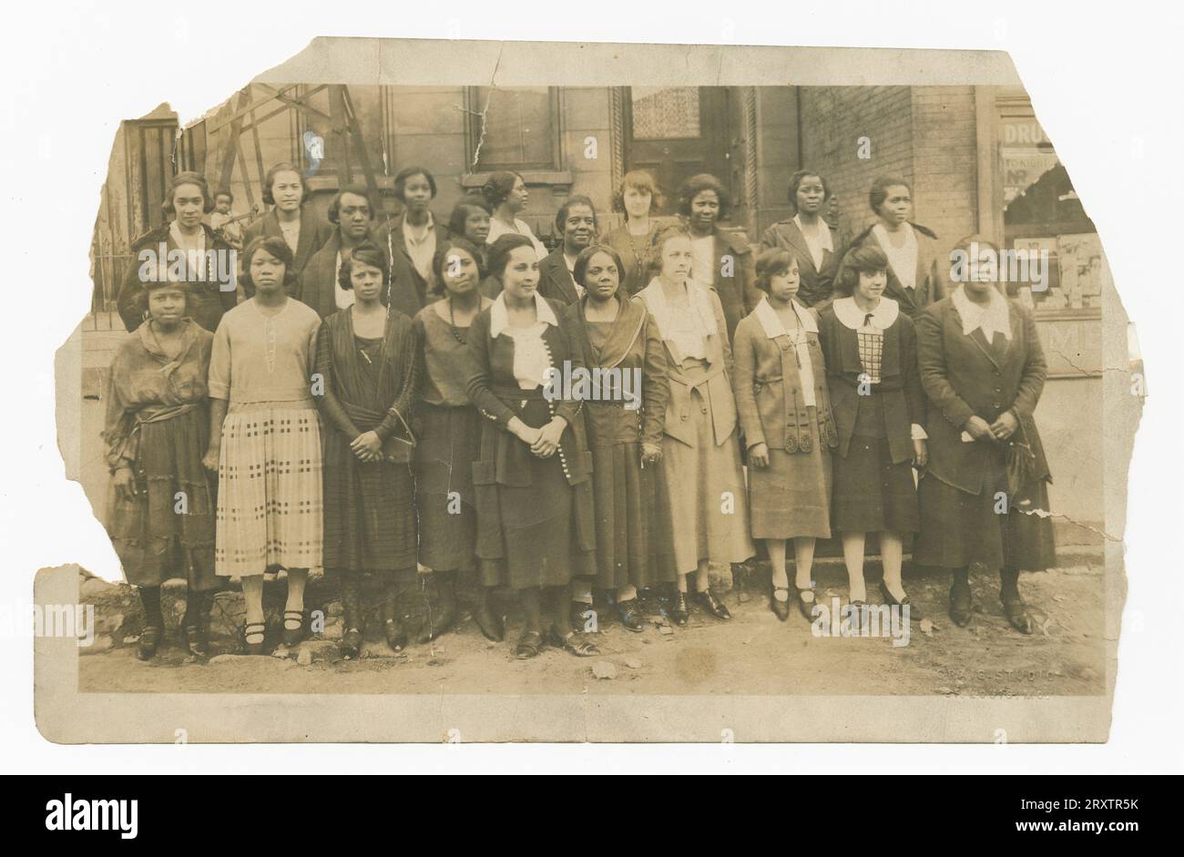 Photograph of tweny-one (21) women and (1) one small child.  The women stand in rows on the steps of a building.  The women wear various different styles and patterns of dress. In the upper right corner of the image is a window with advertisements and products within the window bay.  At the top of the image in the center is a doorway.  There are curtains in the window of the door.  In the upper left corner of the image is some wooden scaffolding.  In the scaffolding is the small child, looking at the camera.  In the lower right corner of the image is an impression of the photographer [CRAIG ST Stock Photo