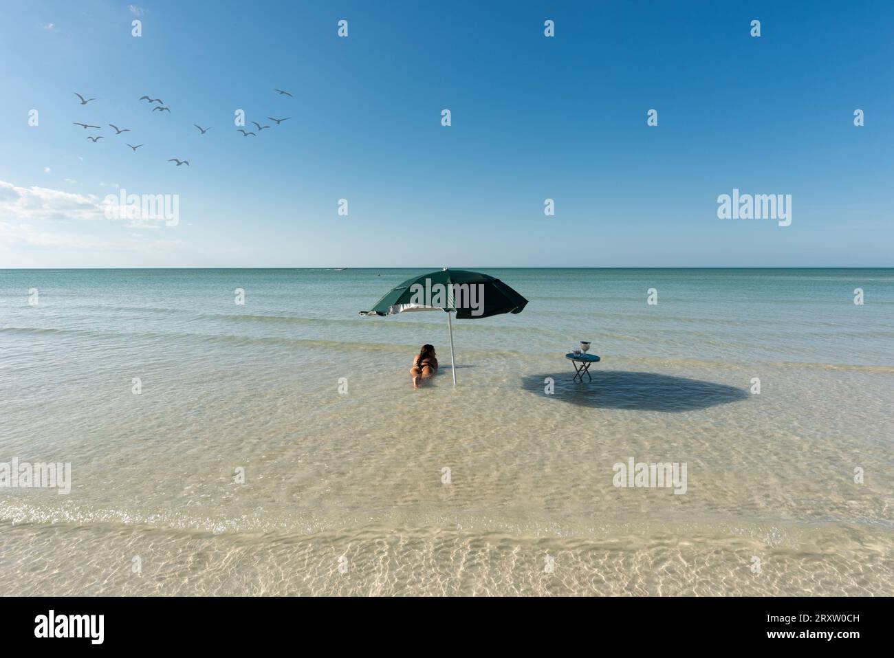 Young woman on vacation as she relaxes in the sea on the shallow sandy bottoms of the Caribbean Sea, next to her a beach umbrella and a small table wi Stock Photo