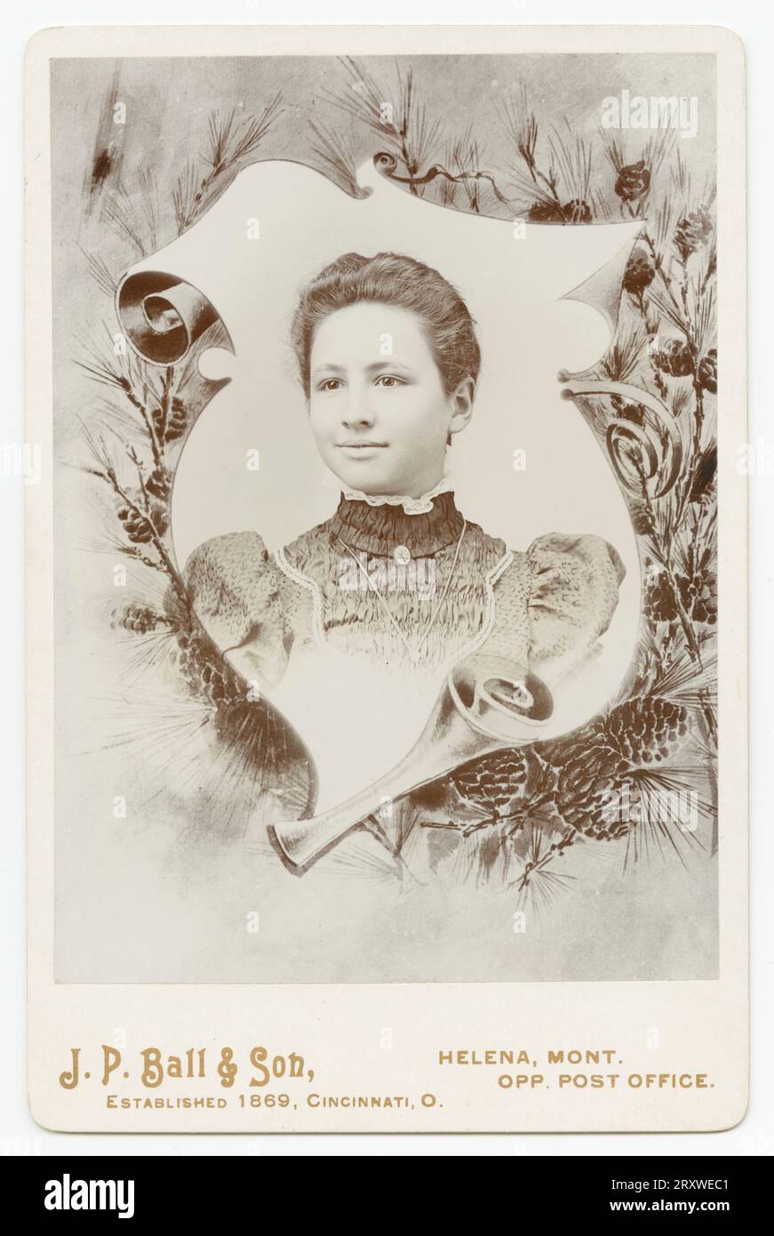 A black-and-white photograph of a woman photographed at the J. P. Ball & Son studio in Helena, MT. The photo is mounted on a light colored cabinet card with the name and location of the photo studio printed on the front along the bottom. The portrait is surrounded by a decorative border of curling scroll paper and pine tree branches. The girl is wearing a patterned dress with ruffled collar and locket. She has dark hair that is pinned back. There is an inscription in pencil on the back of the card. Stock Photo
