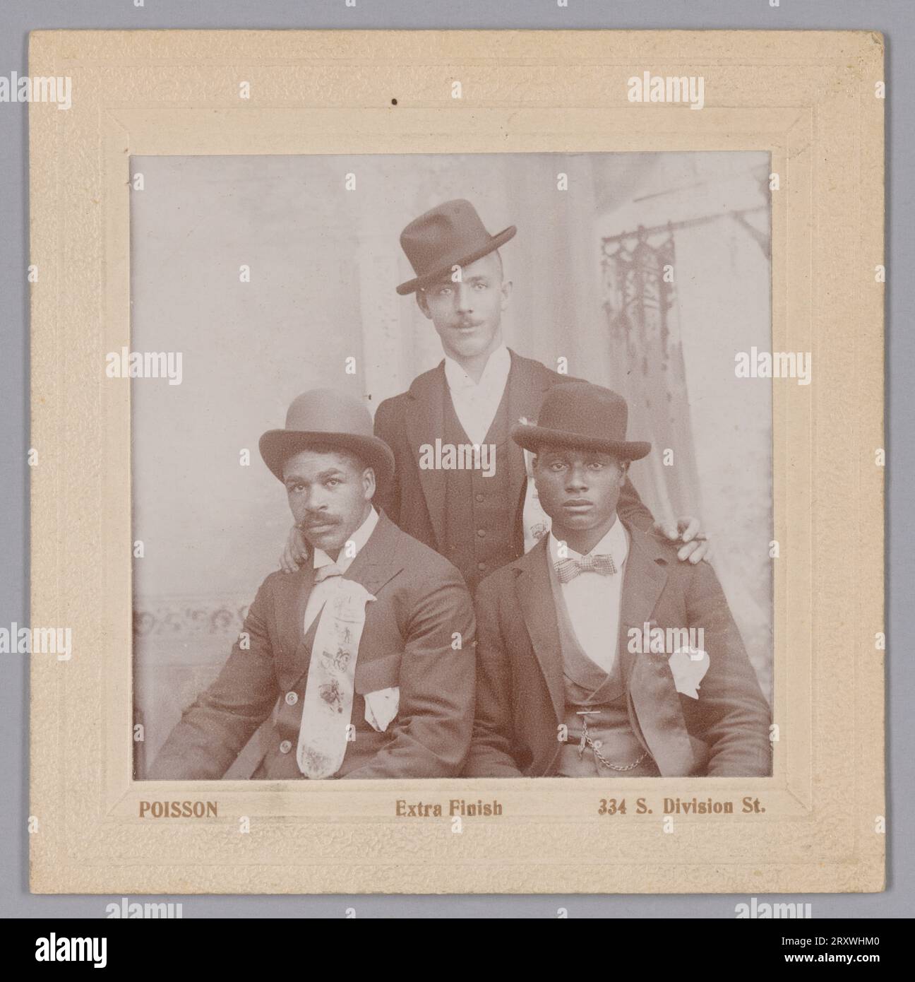This black-and-white photograph by Joseph H. Poisson shows three (3) unidentified men who all wear dark suits, hats, and each have a wide ribbon attached to their proper left jacket lapel. Two men are seated with the third standing behind and between them, with his hands resting on their shoulders. The seated man on the left and the standing man both have light-colored ribbons with symbols printed in a darker color on them, while the man seated on the right has a plain dark-colored ribbon. The man on the right is clean-shaven, while the other two have mustaches. The photograph is mounted to a Stock Photo