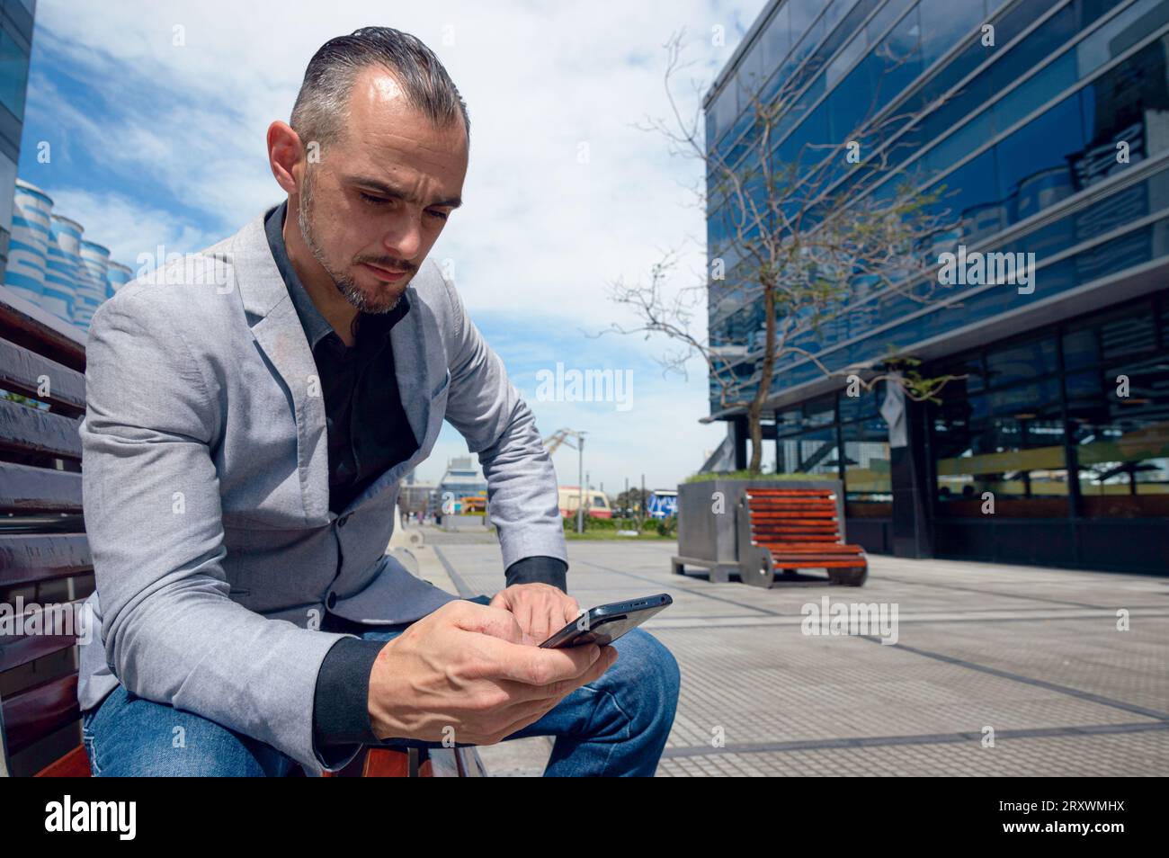 French Caucasian adult businessman with beard wearing stylish clothes, sitting outdoors with commercial buildings around, using phone sending messages Stock Photo
