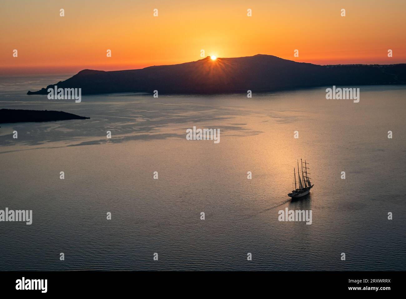 The bay off Thera, Santorini with sun just setting behind the island of Thirasia, golden clear sky and masted sailing ship in middle of bay Stock Photo