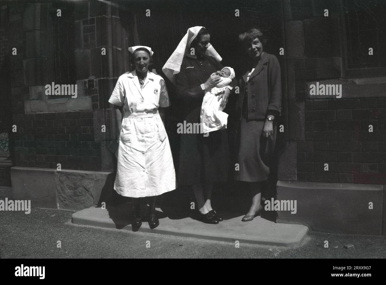 1960s, historical, summertime and at the entrance to a buidling, two uniformed nurses, one holding a new baby, standing outside for a photo beside its new mother, Manchester, England, UK. Stock Photo