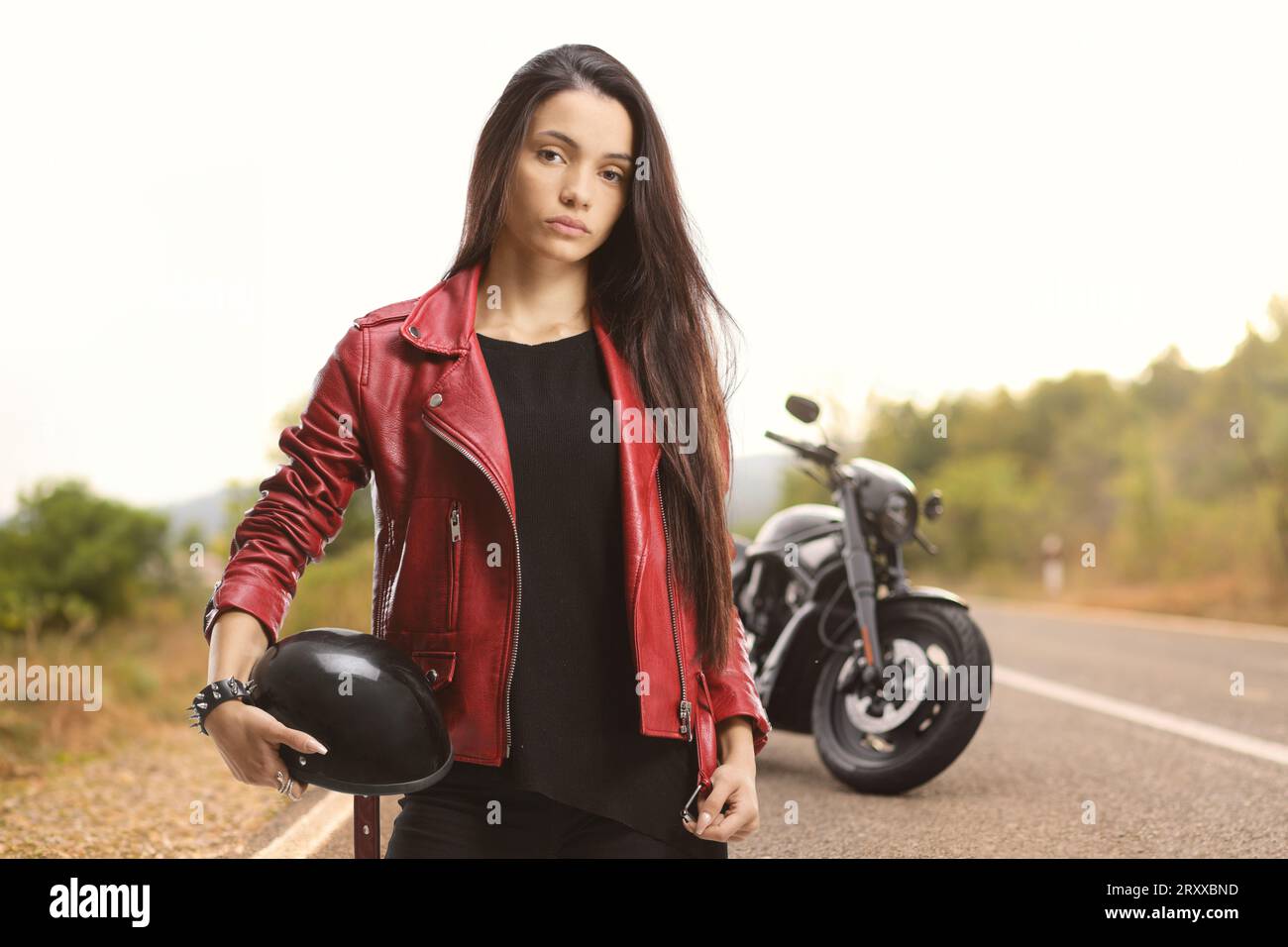 Serious young female holding a motorbike helmet in front of a chopper on the road Stock Photo
