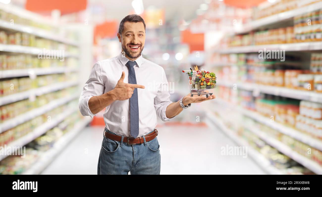 Young smiling man holding a small shopping cart and pointing inside a supermarket Stock Photo