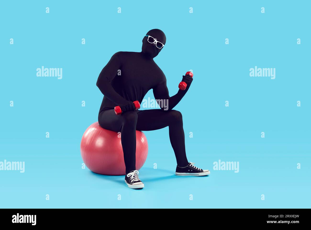 Man disguised in black spandex bodysuit sitting on fit ball and doing exercise with dumbbells Stock Photo