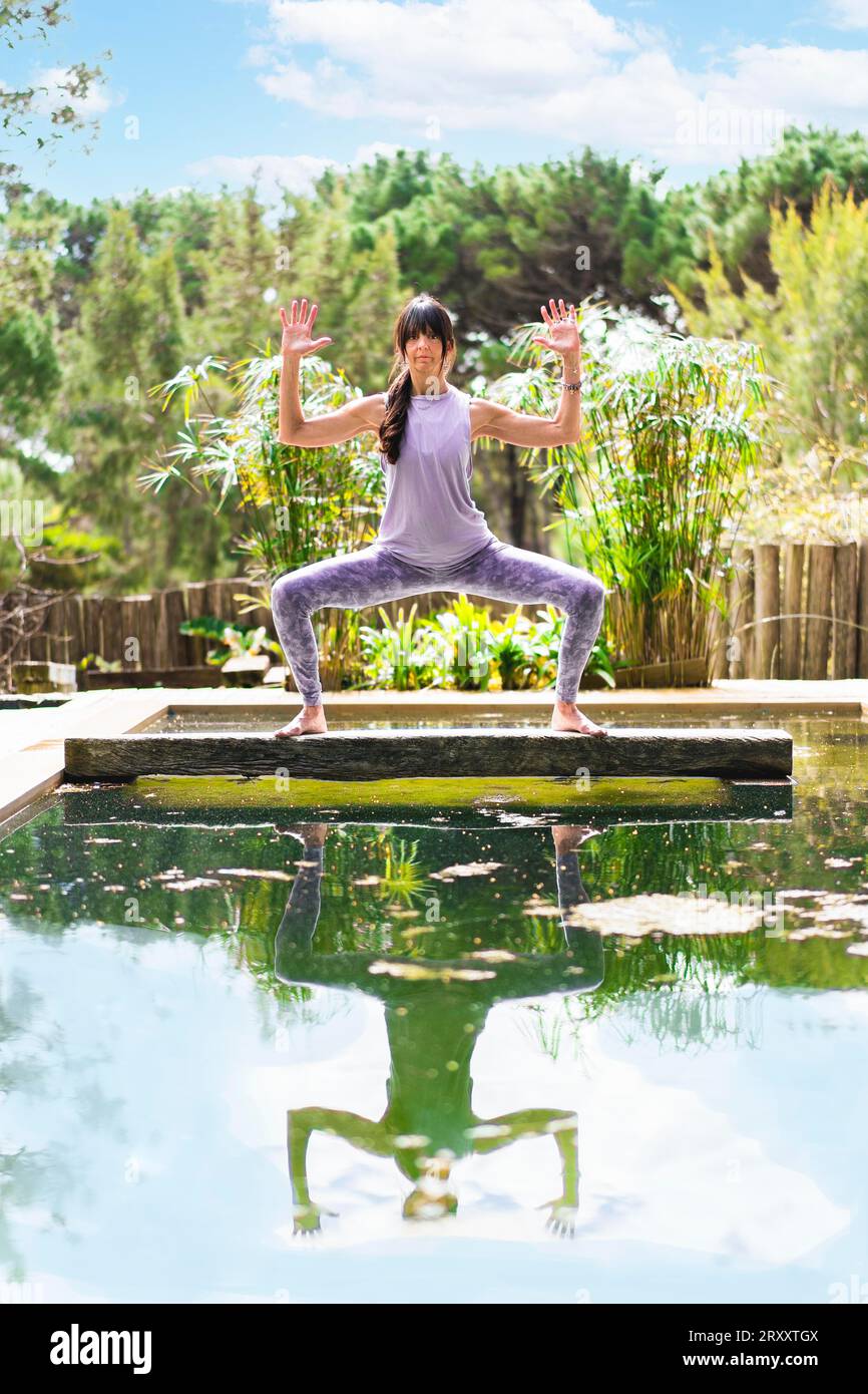 A woman practicing the yoga pose Goddess with Cactus Arms on a tree trunk over a pool, Vertical shot Stock Photo