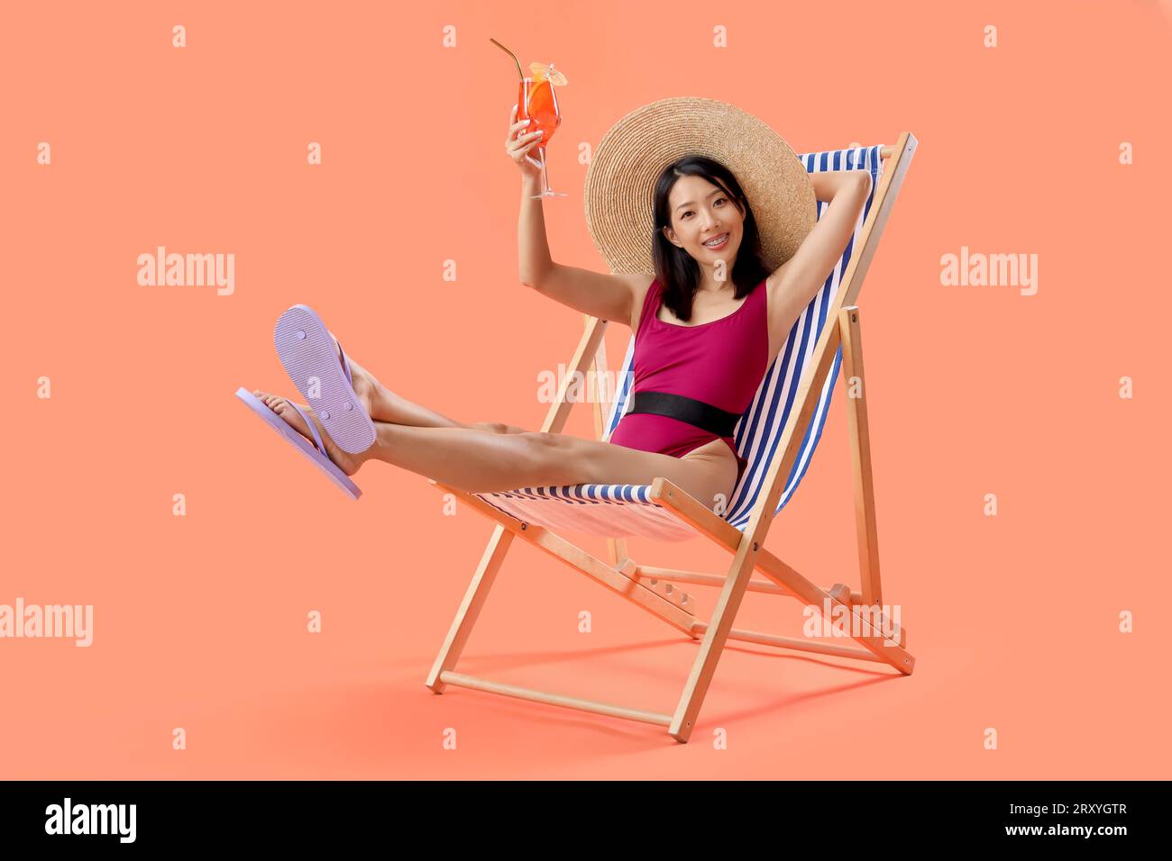 Beautiful young Asian woman in swimsuit with tasty aperol spritz sitting on deckchair against orange background Stock Photo