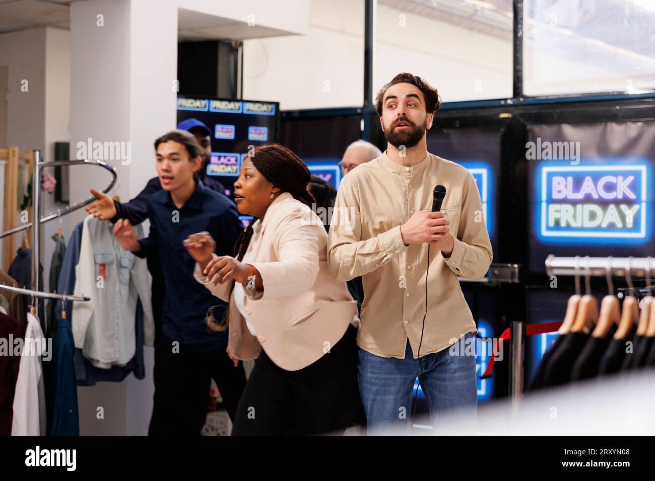 Shocked man video TV news reporter live broadcasting about Black Friday shopping madness, standing with microphone in crowded store. Crazy frenzy crowd running to get best deals into clothes shop Stock Photo