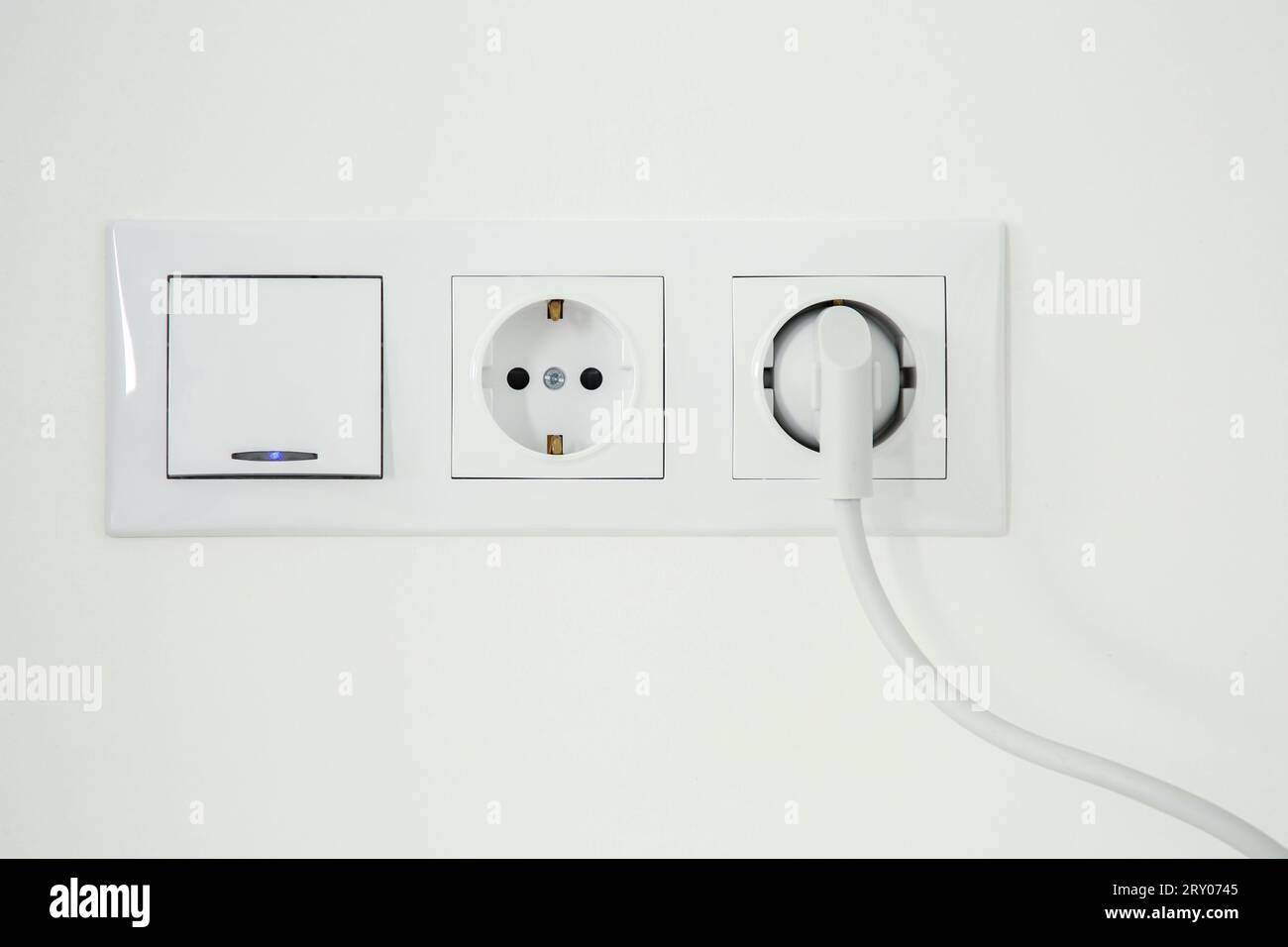 Power sockets with inserted plug and light switch on white wall indoors