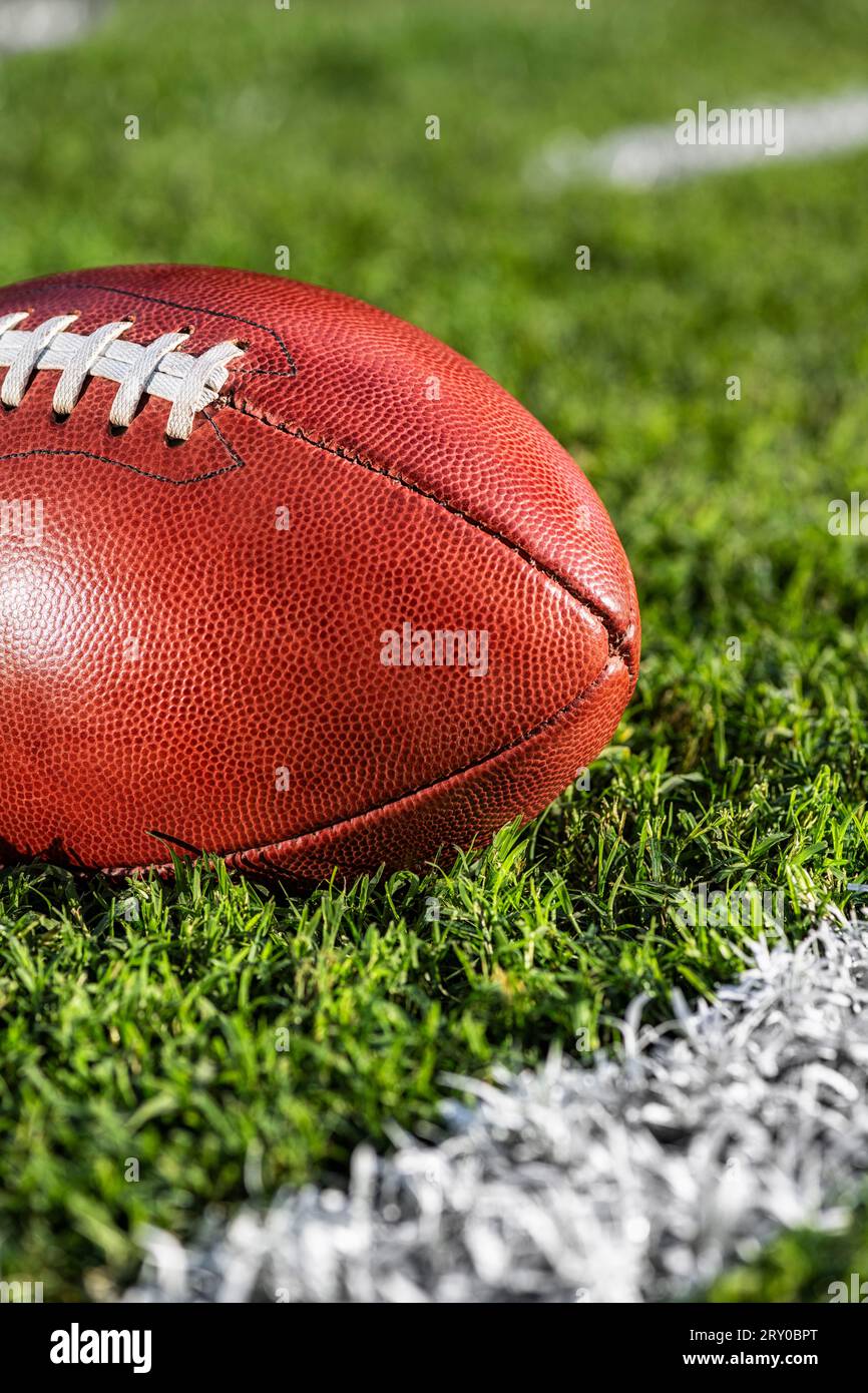 A low angle close-up view of a leather American Football sitting in the grass next to a white yard line with hash marks in the background. Stock Photo