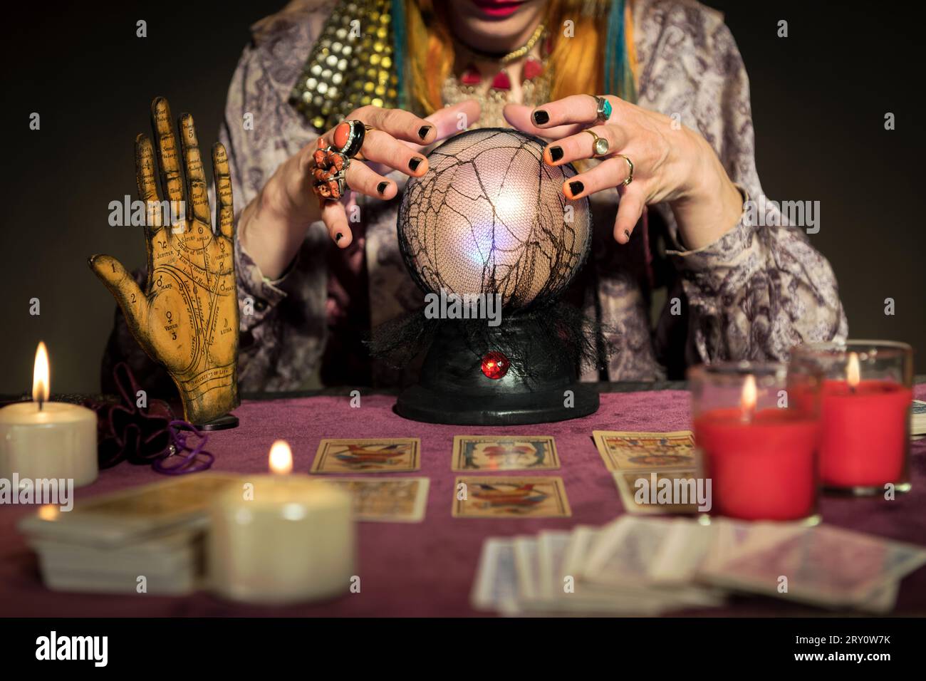 Crop anonymous fortune teller female looking down while sitting at table with hovering fingers on crystal ball burning candles blurred cards and palmi Stock Photo