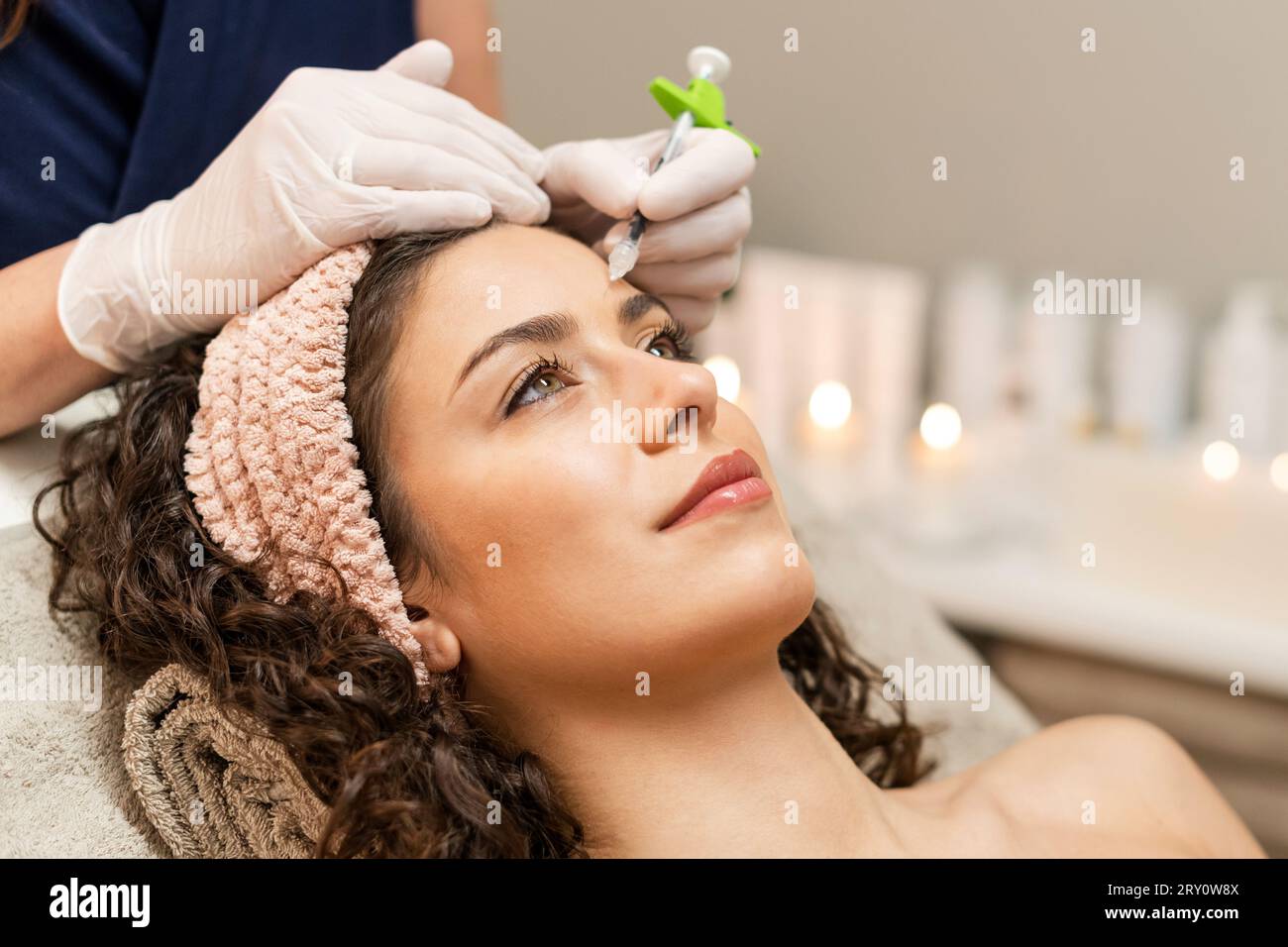 From above of curly female client looking away while unrecognizable cosmetologist injecting anti age filler into forehead in beauty center Stock Photo