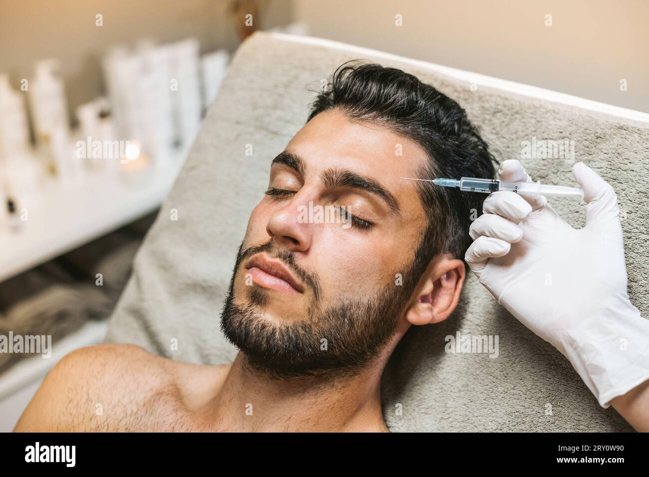 High angle of bearded male customer closing eyes while crop beautician injecting botox into forehead with syringe in beauty salon Stock Photo